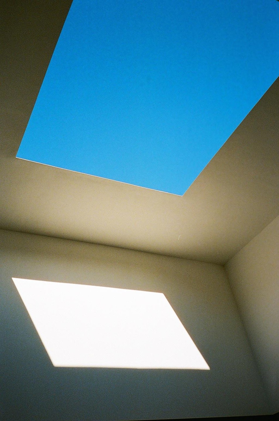 A rectangular skylight shows a clear blue sky, and a parallelogram of sunlight illuminates the wall below.