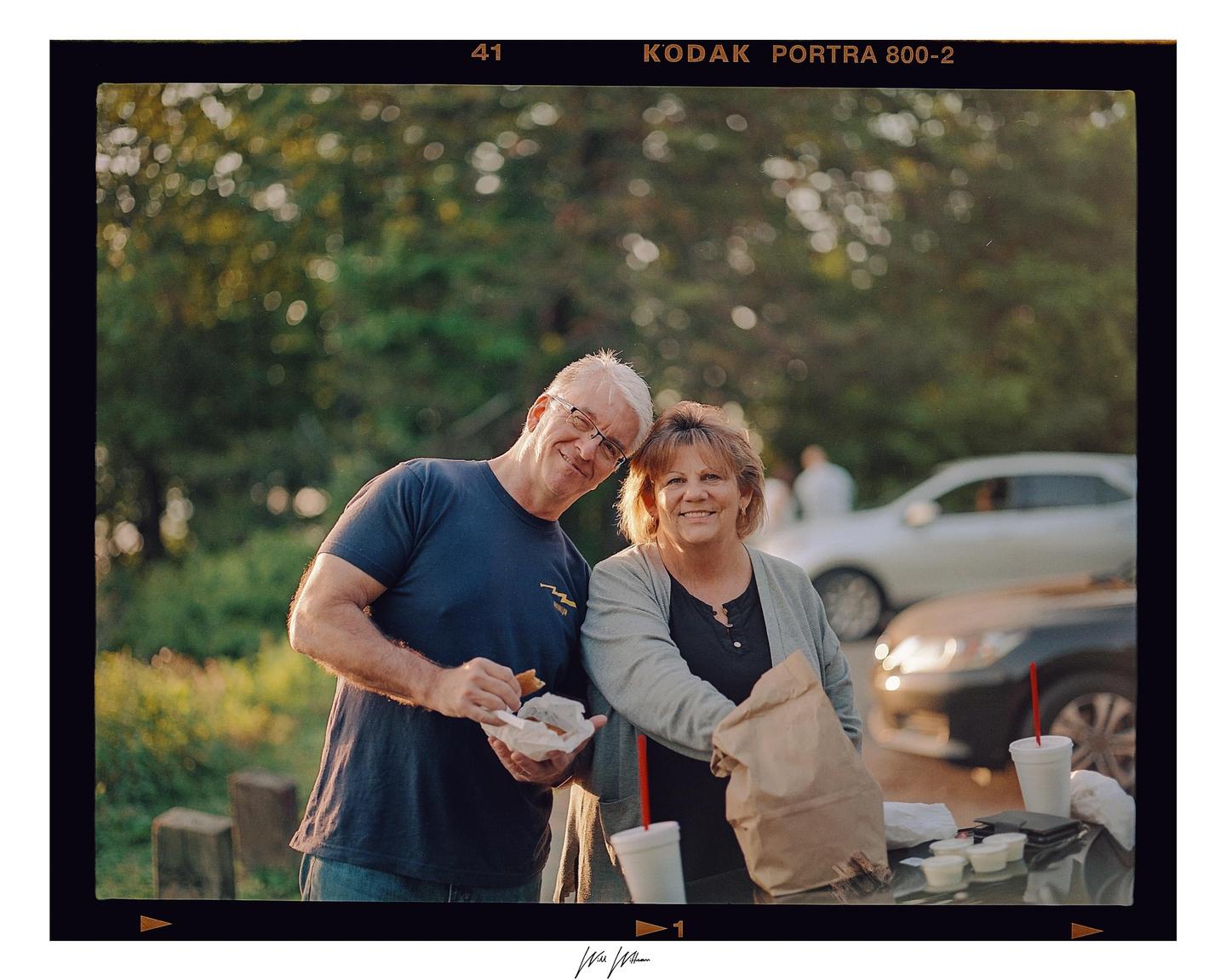 A man and a woman smile at the camera while holding food and drinks outdoors.