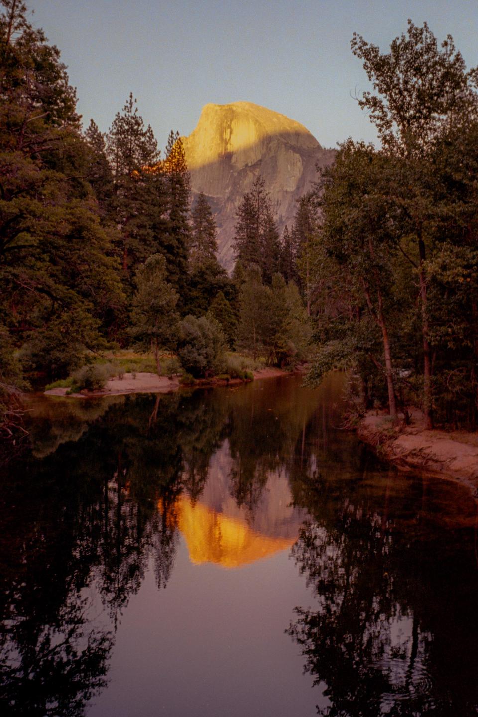 A mountain with a sunlit peak is reflected in a body of water surrounded by trees.