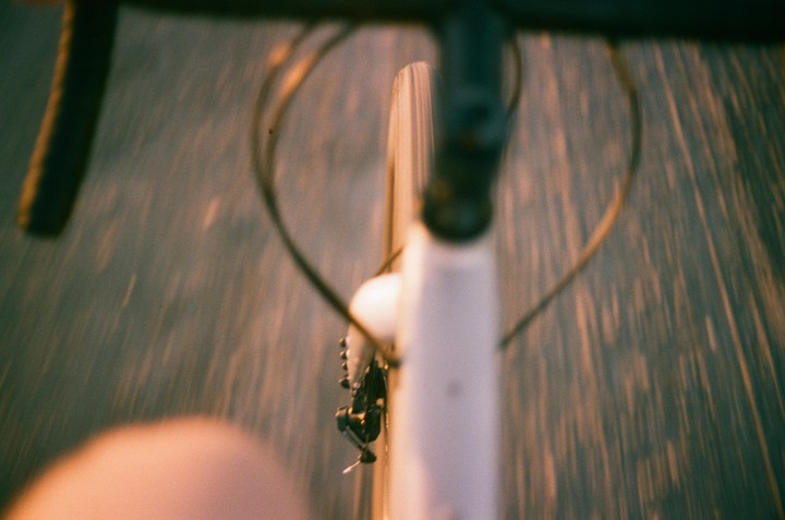 A bicycle handlebar and front wheel are visible from a first-person perspective, with the road blurred beneath.