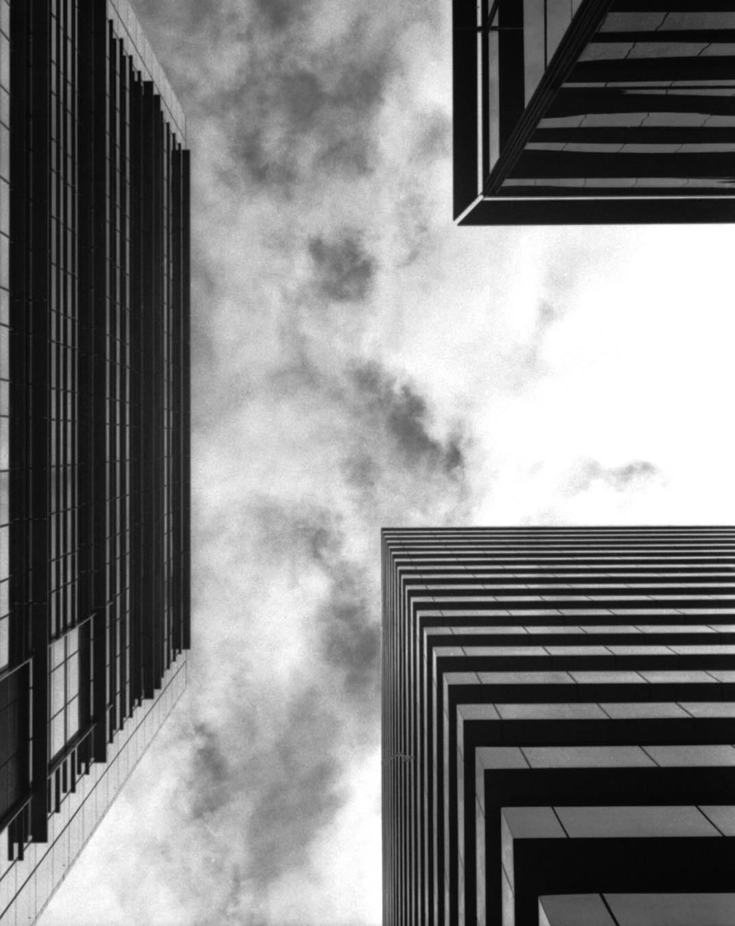 Three modern buildings are viewed from below, with clouds in the sky between them.