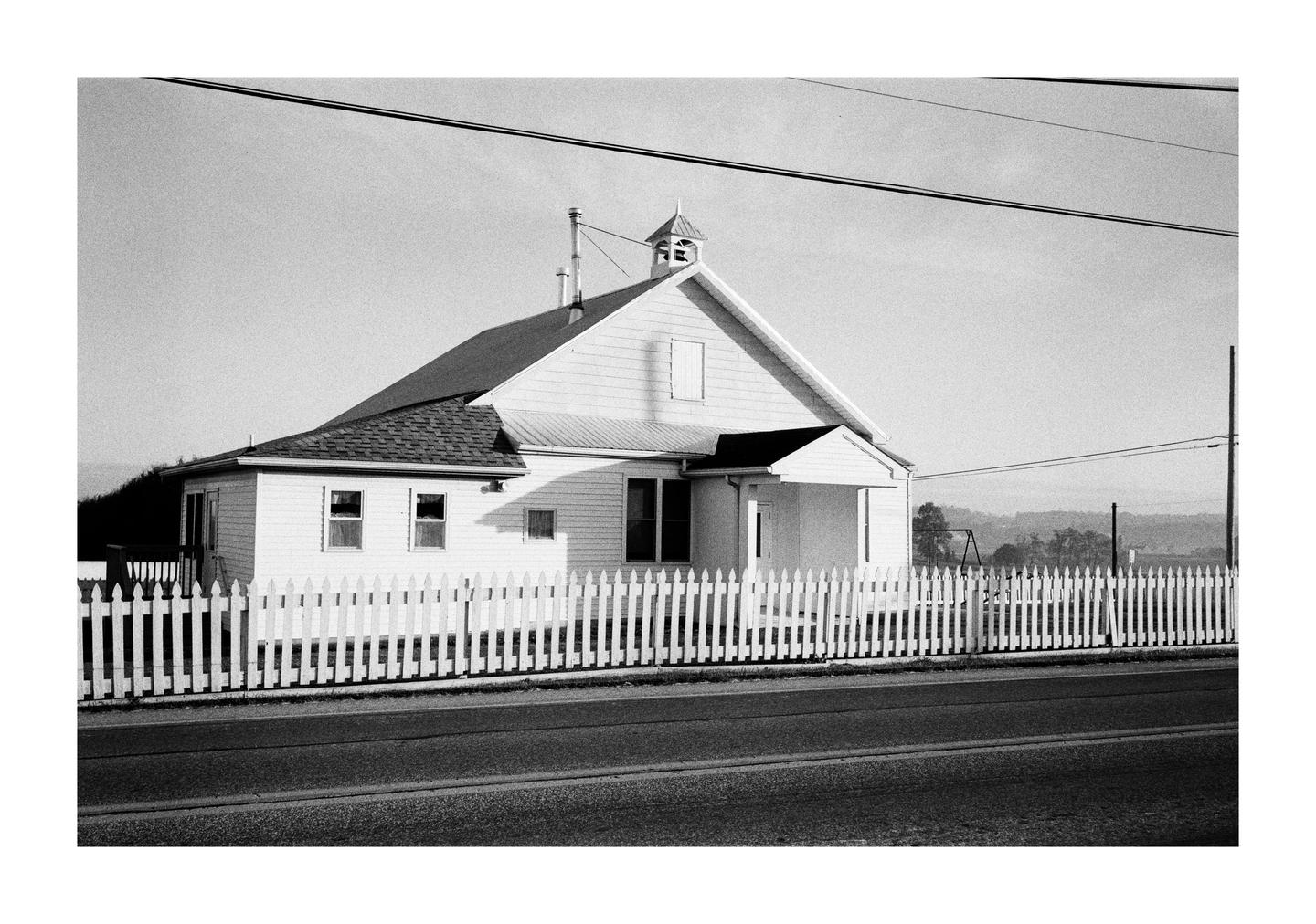A white house with a steeple and a picket fence stands beside a road under a clear sky.