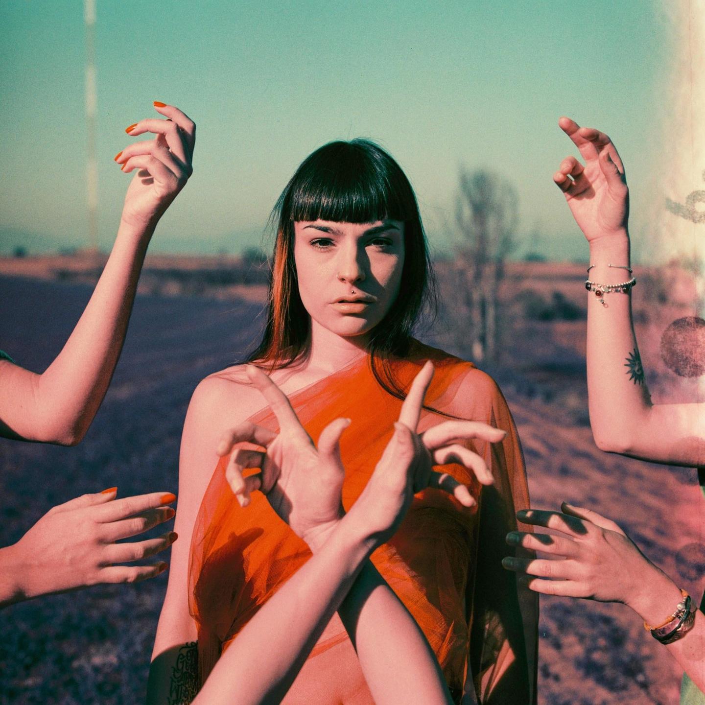 A woman with a septum piercing and orange hair streaks is surrounded by multiple hands in a field.
