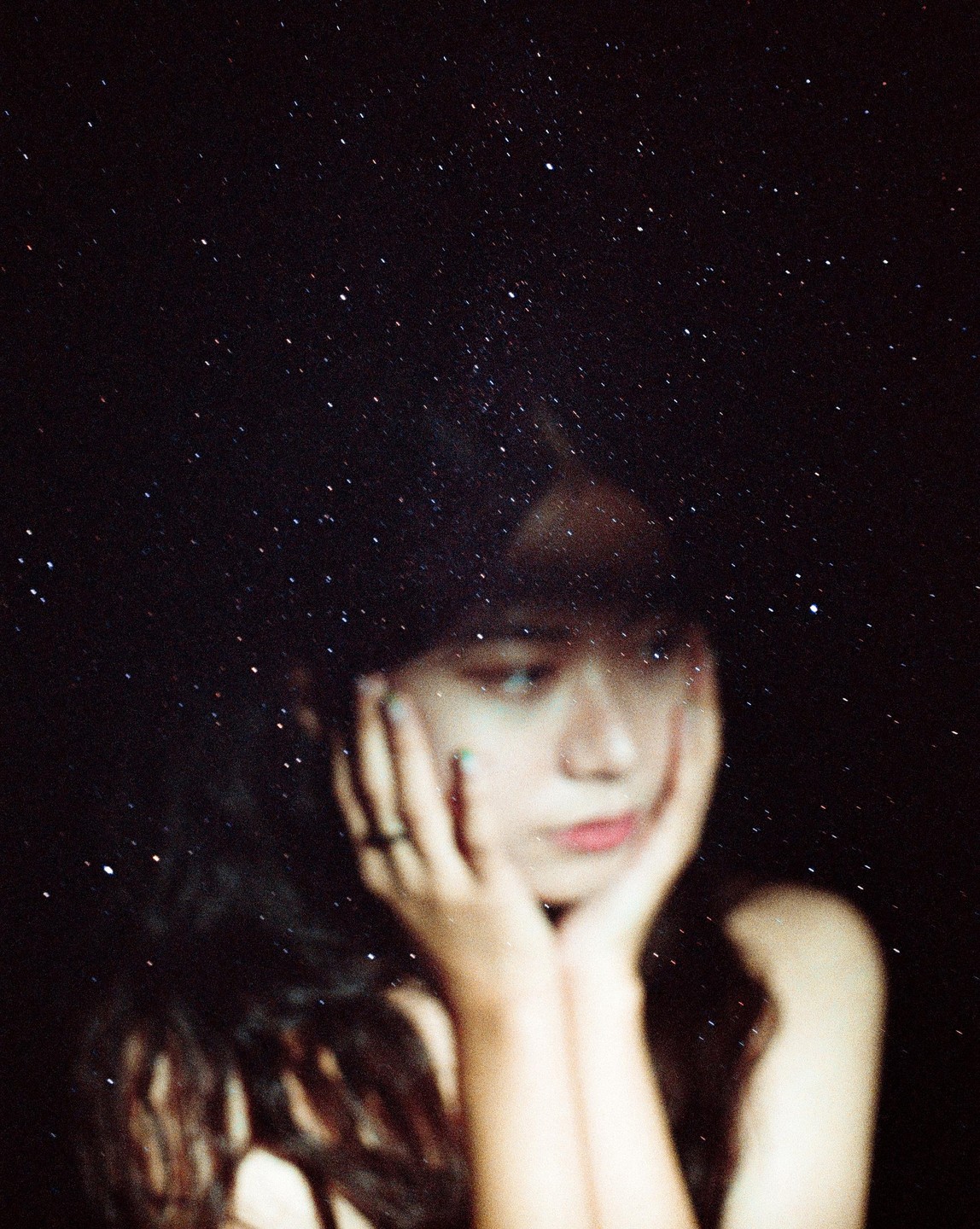 A woman with long hair rests her chin on her hands, with a starry sky superimposed over her head and shoulders.