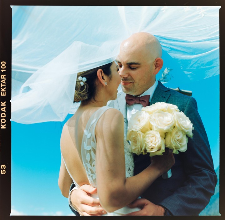 A couple embraces, with the woman holding a bouquet of white roses, under a veil against a bright blue sky.