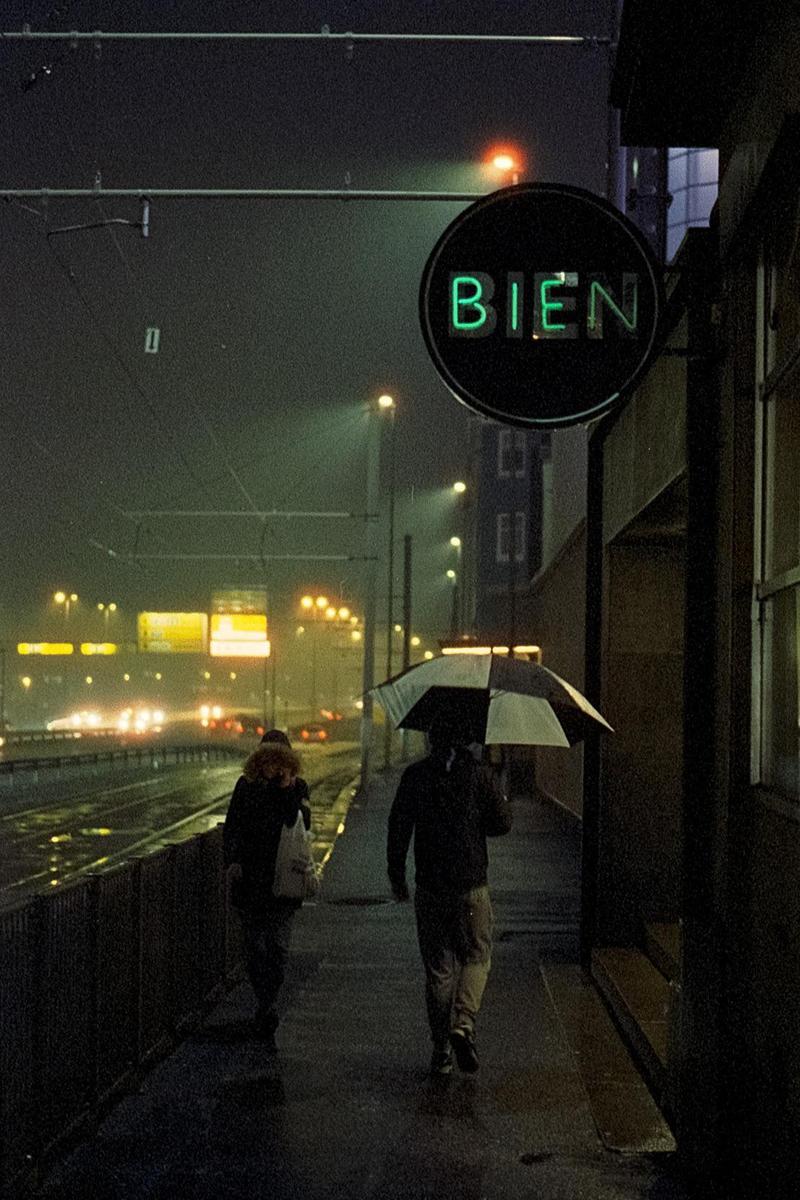 Two people walk on a wet street at night under a neon sign that reads "BIEN" while cars drive by in the background.