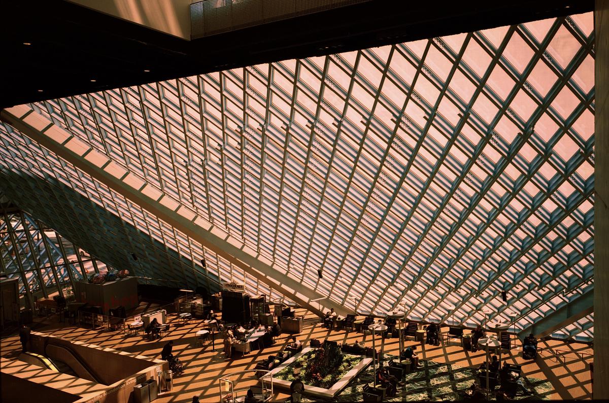 The interior of a modern building features a large glass ceiling and people seated at tables on the floor below.