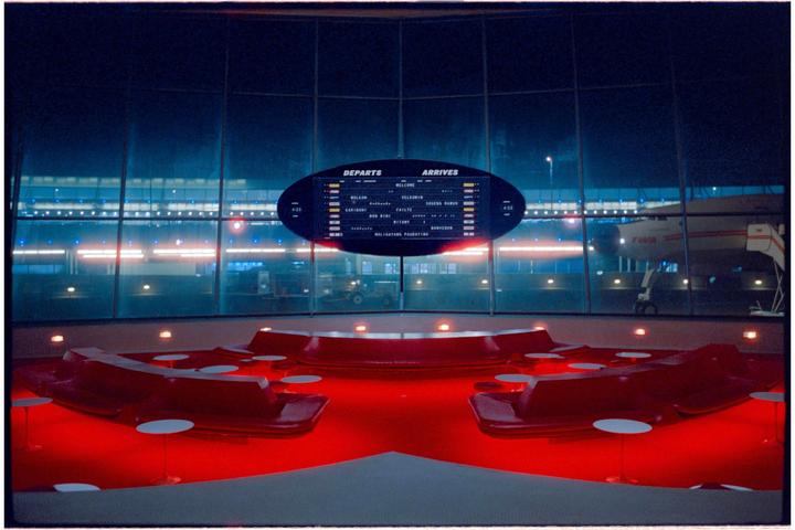 A departure and arrival board is visible through a large window overlooking an airport tarmac at night, with red seating in the foreground.