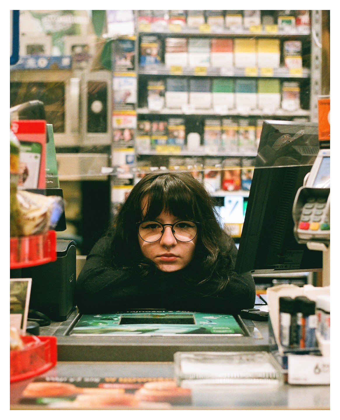 A woman with dark hair and glasses rests her head on her arms at a checkout counter.
