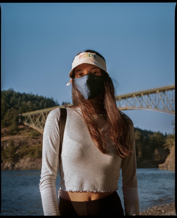 A woman wearing a face mask and a visor with the word "PRIDE" on it stands in front of a body of water and a bridge.