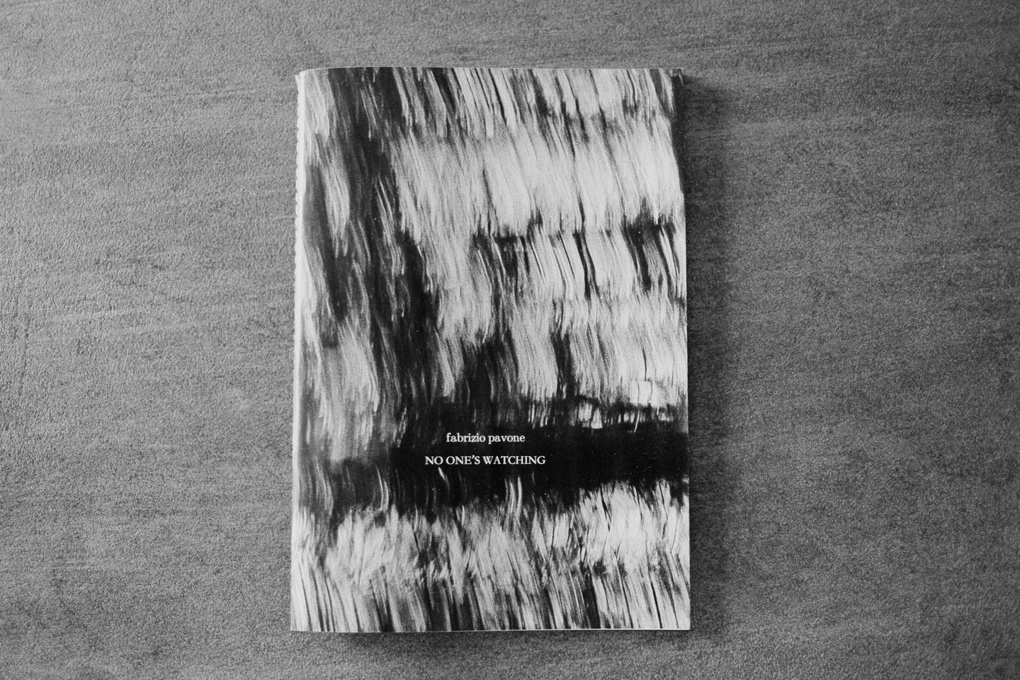 A black and white book with a textured cover titled "No One's Watching" is displayed on a gray surface.