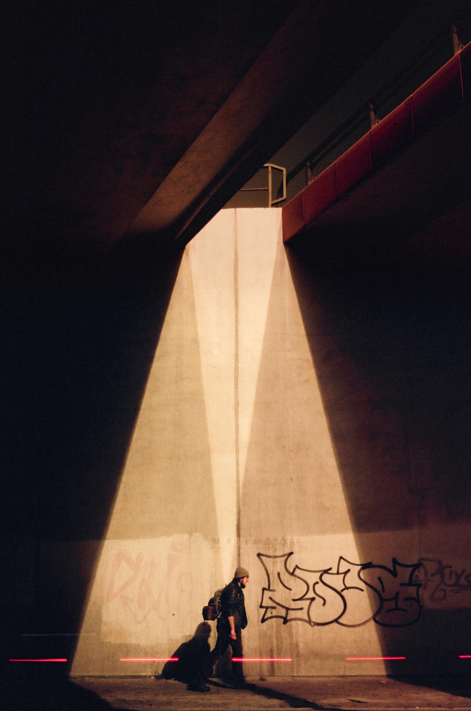 A man with a backpack walks under a bridge with graffiti on the wall, illuminated by light and red laser lines.