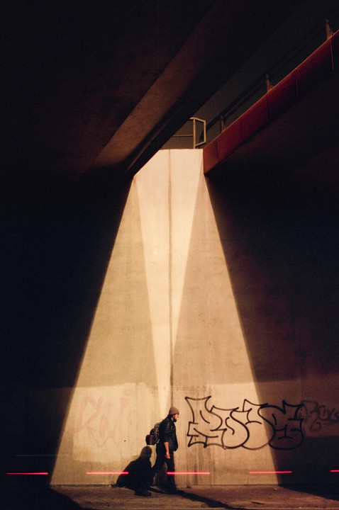 A man with a backpack walks under a bridge with graffiti on the wall, illuminated by light and red laser lines.