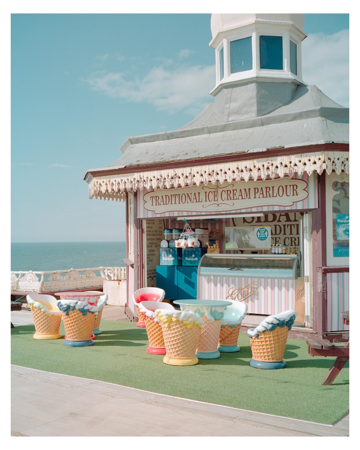 An ice cream parlor with ice cream cone chairs and tables sits on a pier overlooking the ocean.