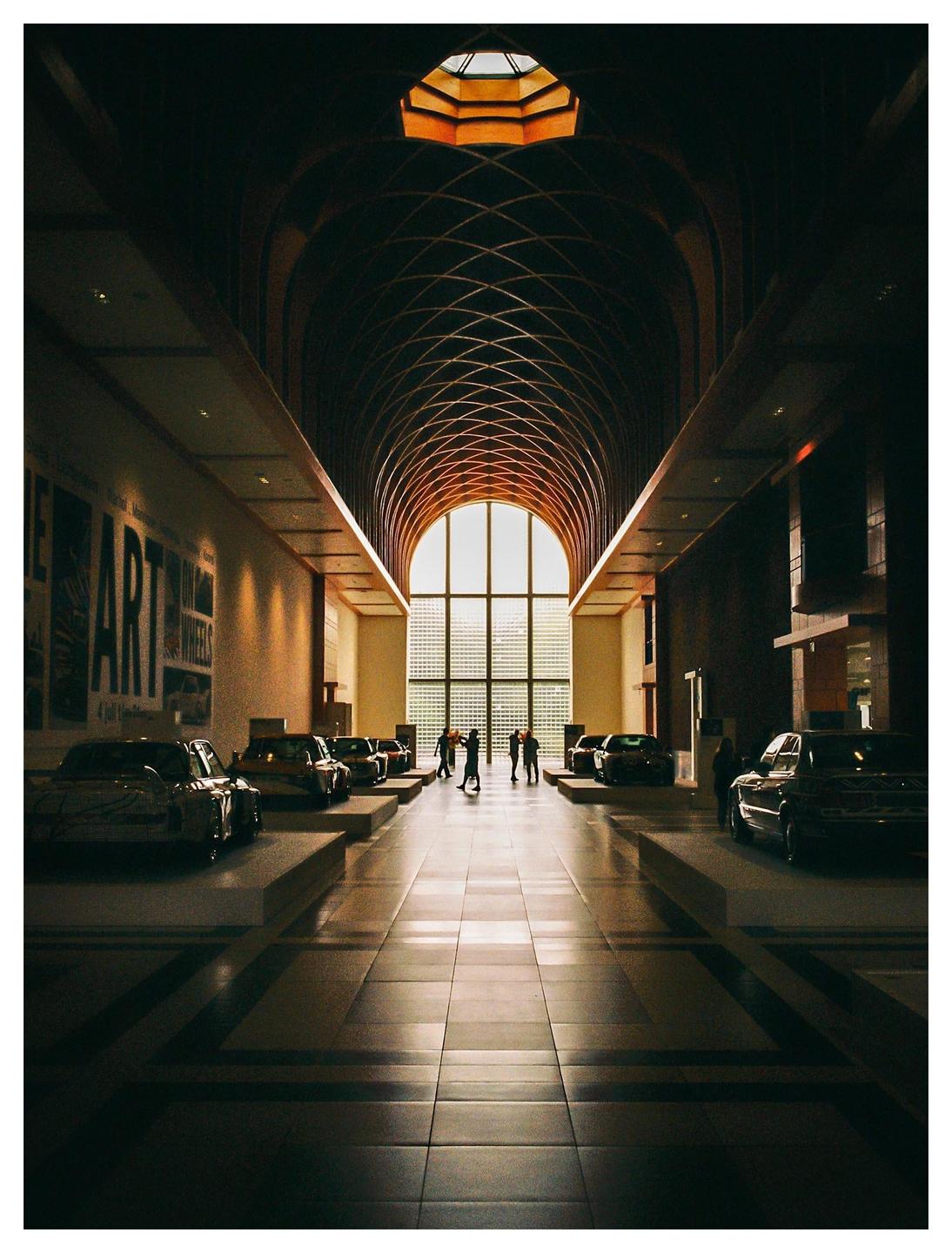 A long hall with a vaulted ceiling and cars on display leads to a large arched window with people standing in front of it.