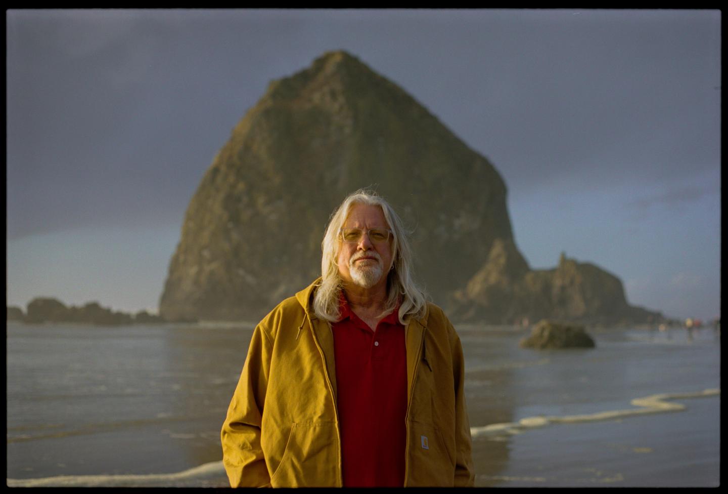 A man with long white hair and a beard stands on a beach with a large rock formation in the background.