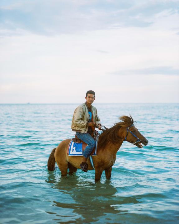 A man rides a horse in the ocean with a cloudy sky in the background.