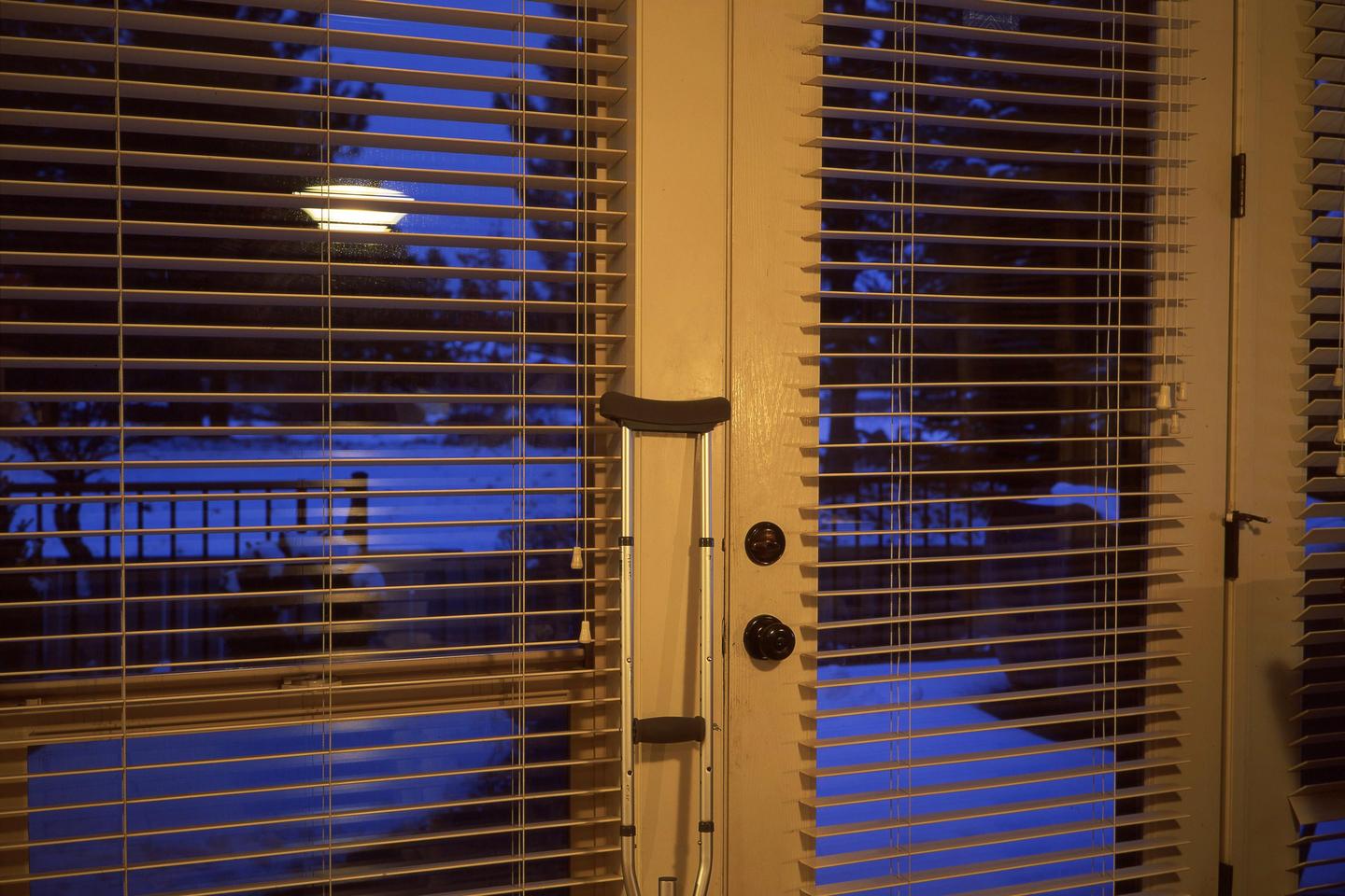 A crutch leans against a door with blinds, revealing a blue outdoor scene with snow and a street light.