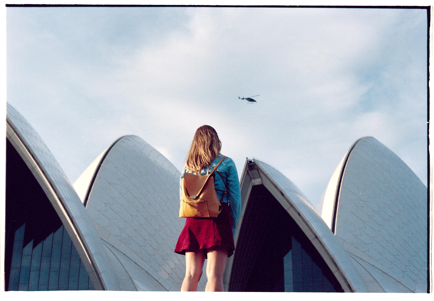 A woman with a backpack stands in front of the Sydney Opera House, looking up at a helicopter in the sky.