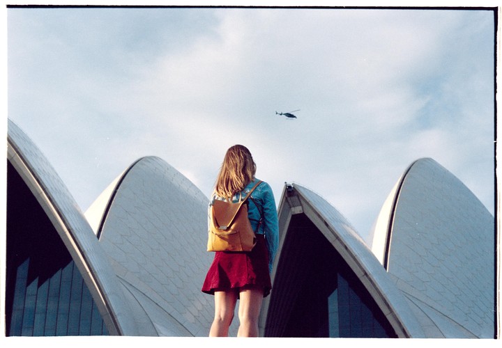 A woman with a backpack stands in front of the Sydney Opera House, looking up at a helicopter in the sky.