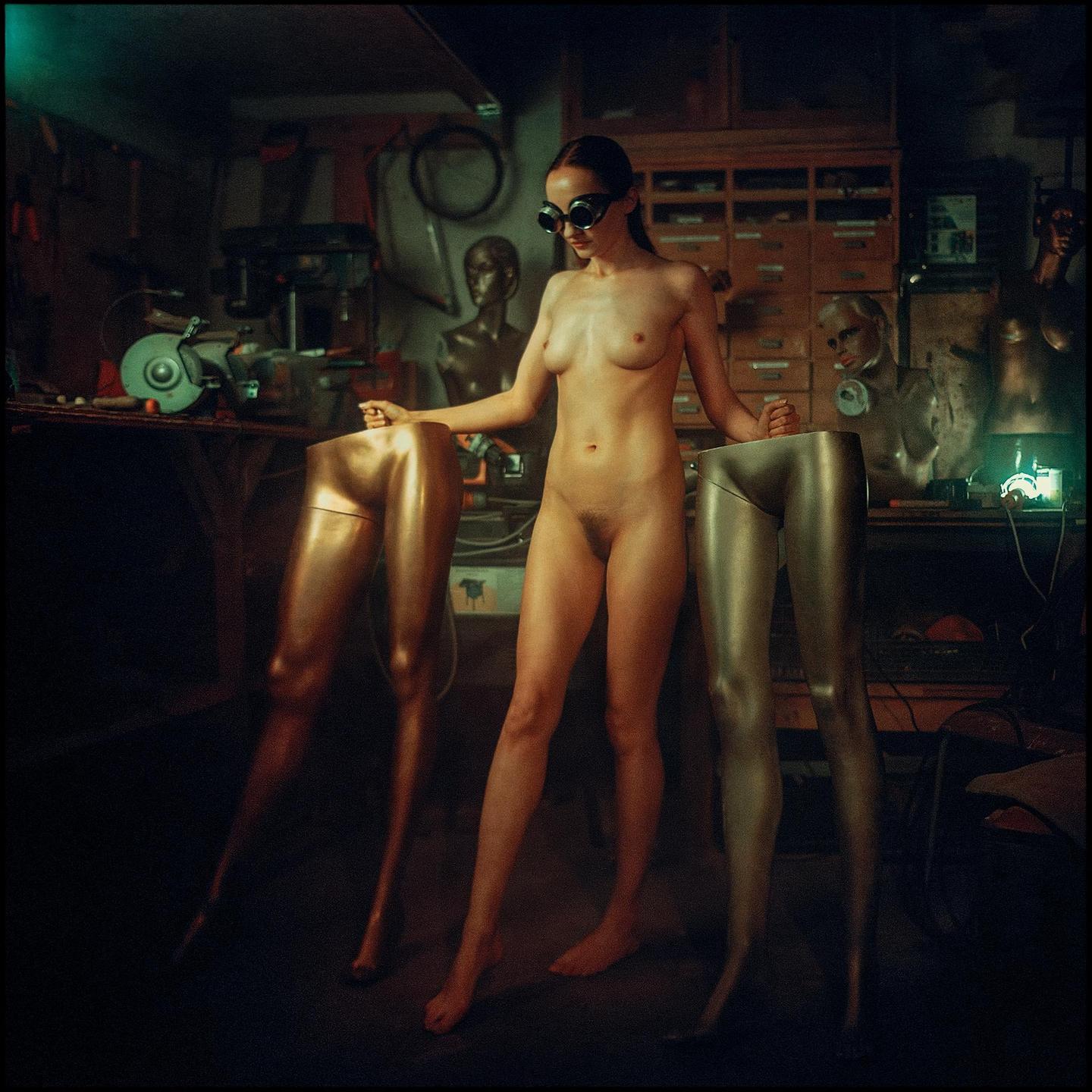 A nude woman wearing goggles stands between two mannequin legs in a workshop.