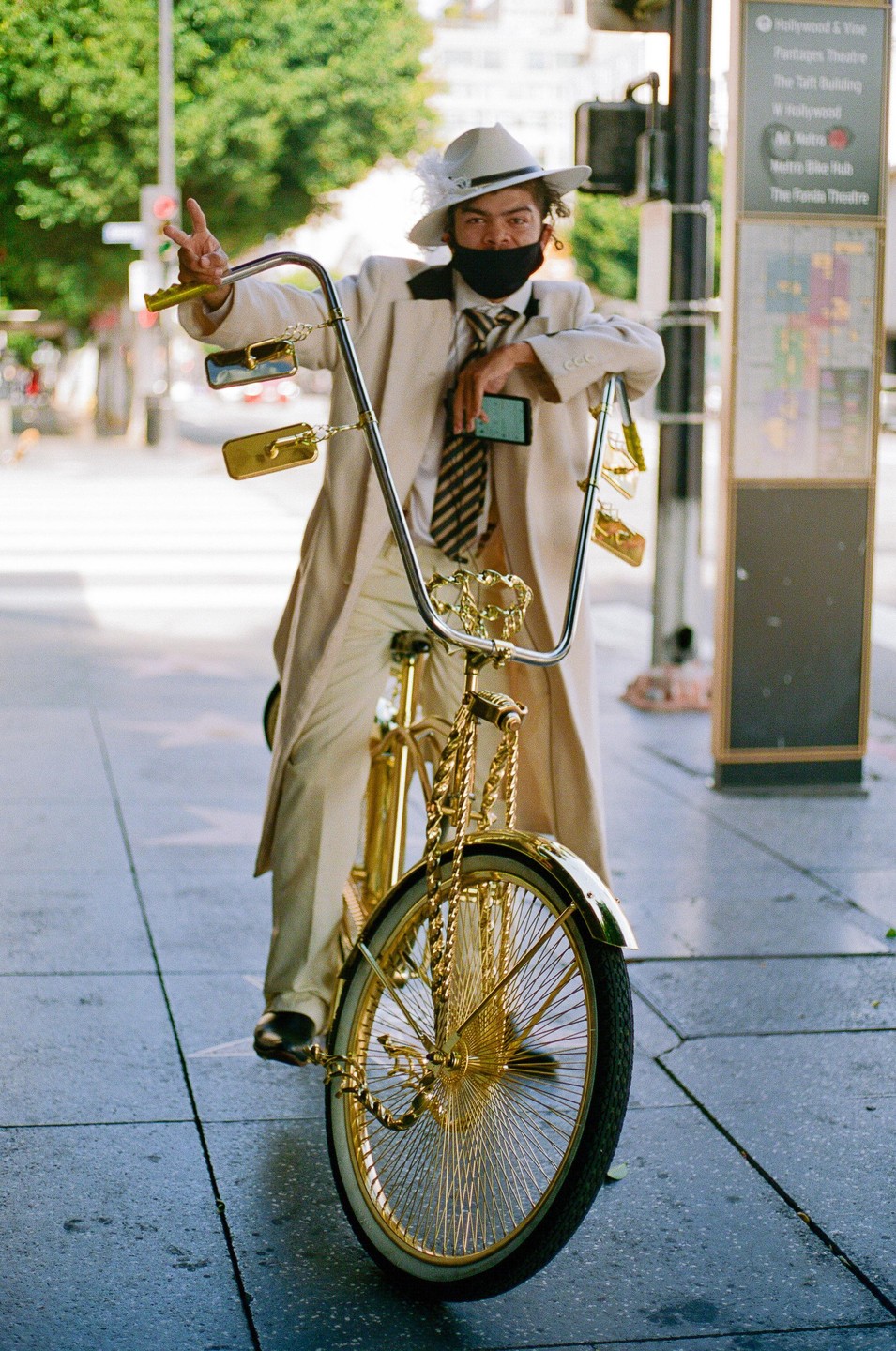 A man in a suit and hat sits on a gold bicycle on a city street, making a peace sign.