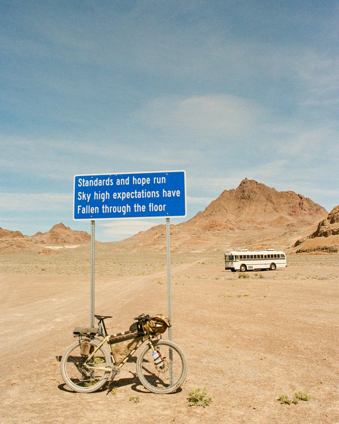A bicycle is parked next to a blue sign with text in a desert landscape with mountains and a bus in the distance.