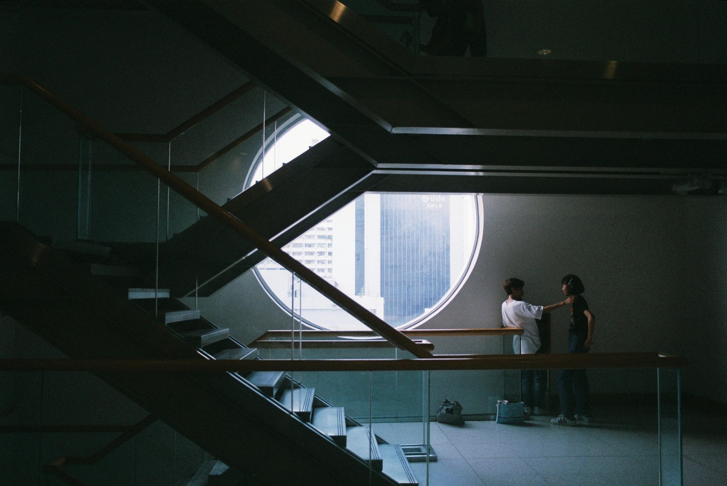 Two people stand near a large circular window overlooking a city, with a staircase in the foreground.