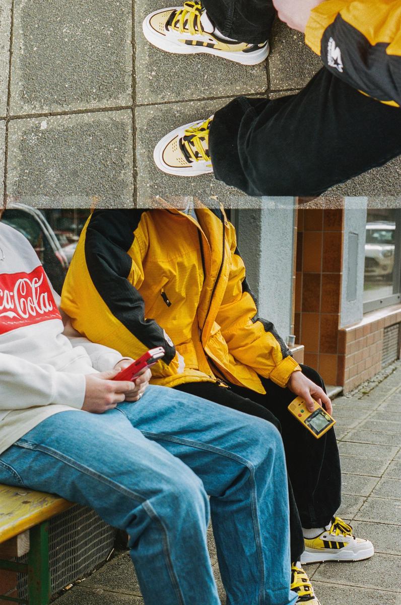 Two people sit on a bench, one holding a red flip phone and the other a yellow handheld game console.