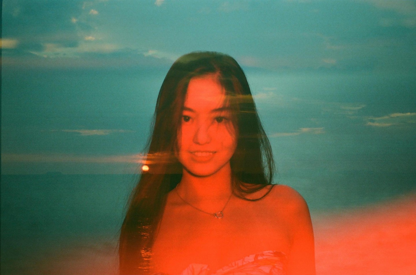 A woman with long hair smiles at the viewer, with a sunset over the ocean in the background.