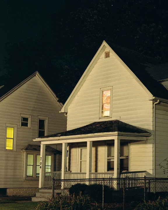 Two houses are illuminated at night, with one showing a figure in an upstairs window.