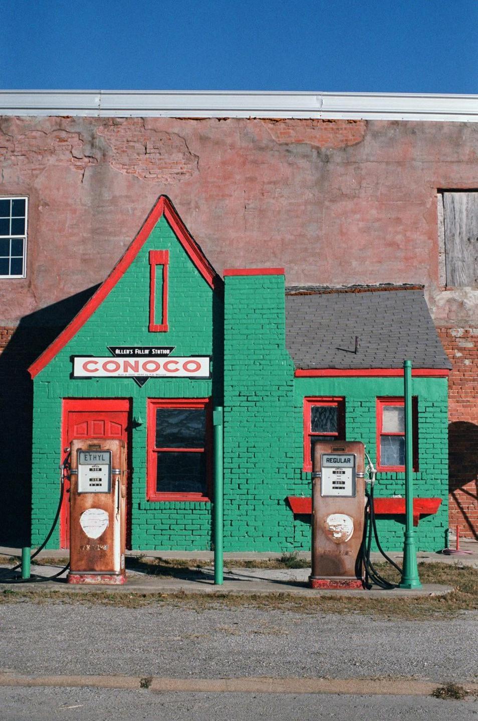 A small, green and red brick building with two vintage gas pumps sits in front of a larger brick building under a clear blue sky.