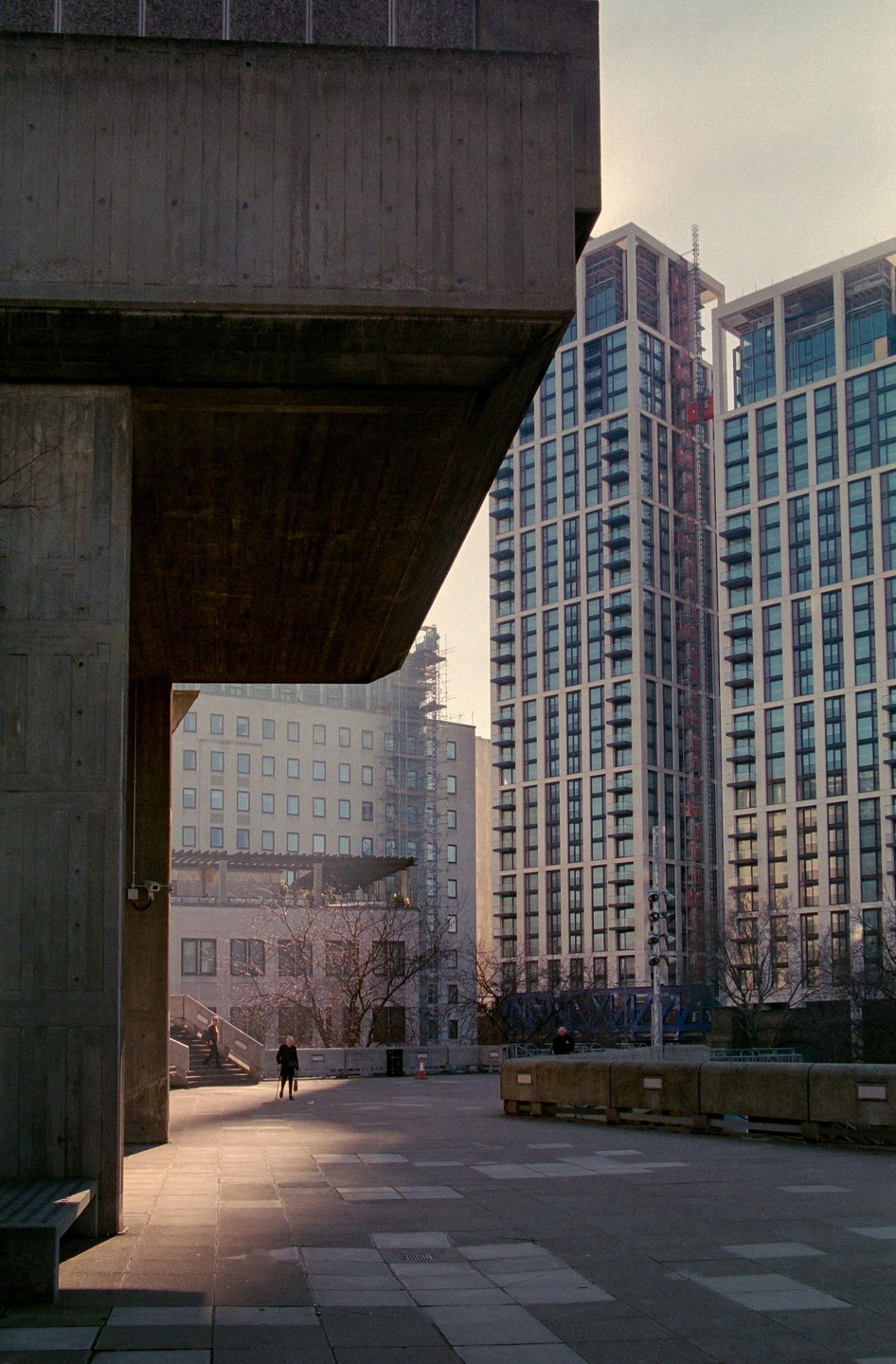 A person walks across a sunlit concrete plaza surrounded by tall buildings and brutalist architecture.