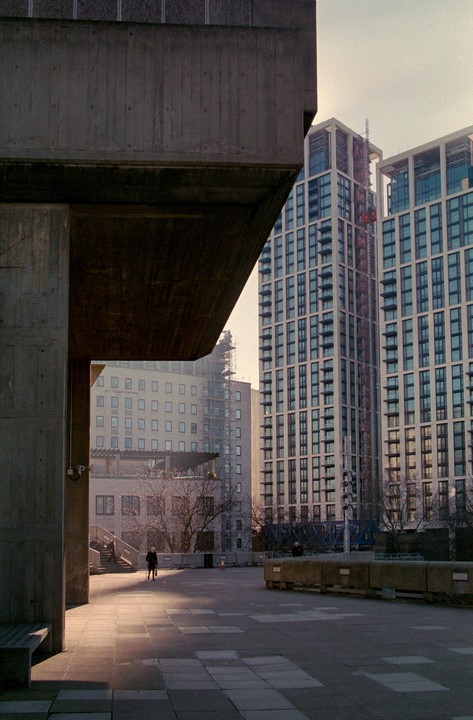 A person walks across a sunlit concrete plaza surrounded by tall buildings and brutalist architecture.