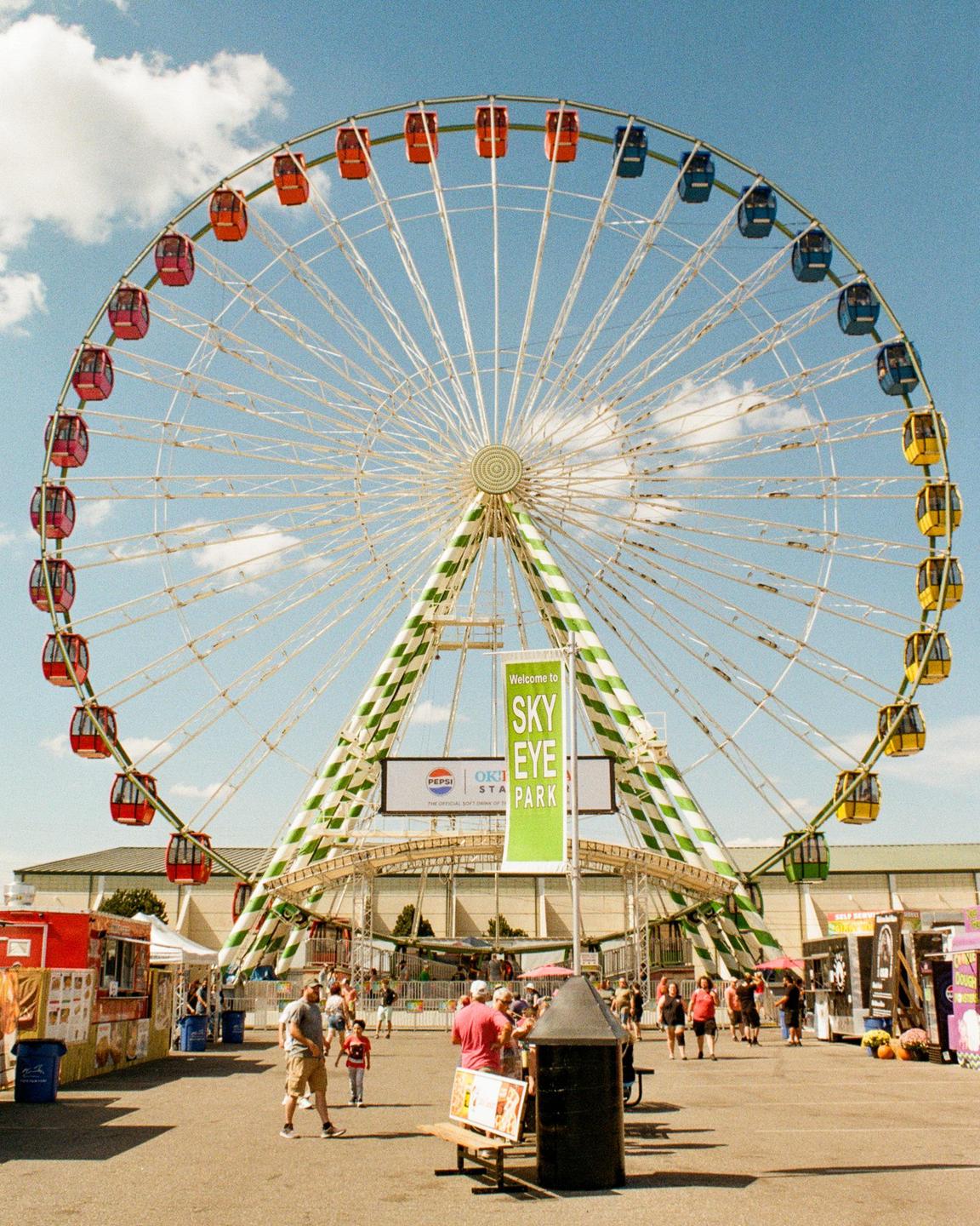 A large Ferris wheel with colorful gondolas stands tall against a blue sky with scattered clouds, overlooking a fairground bustling with people and food stalls.