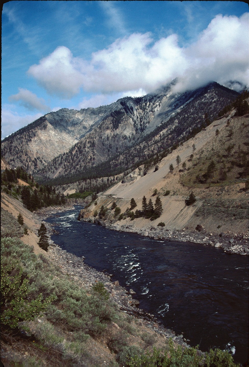 A river flows through a valley with mountains on either side, some of which are covered in clouds.