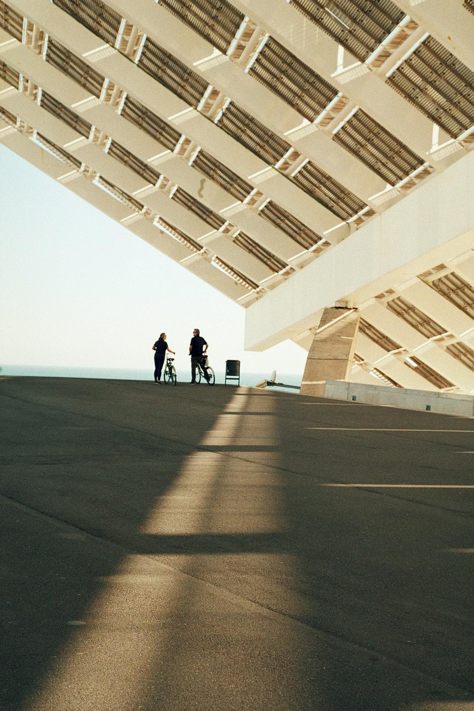Two people stand with their bicycles under a large, modern, angled roof structure, with the ocean visible in the distance.