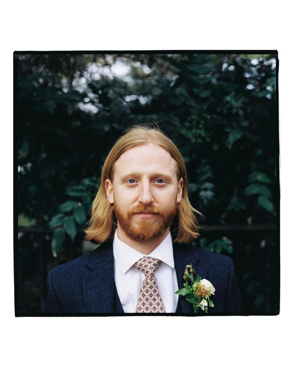 A man with long blond hair and a red beard wears a suit and tie with a boutonniere.