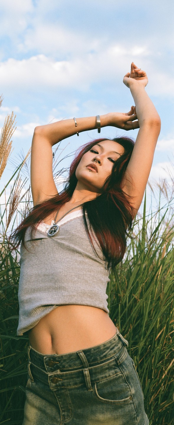 A woman with red hair and a nose ring stands in a field with her arms raised.