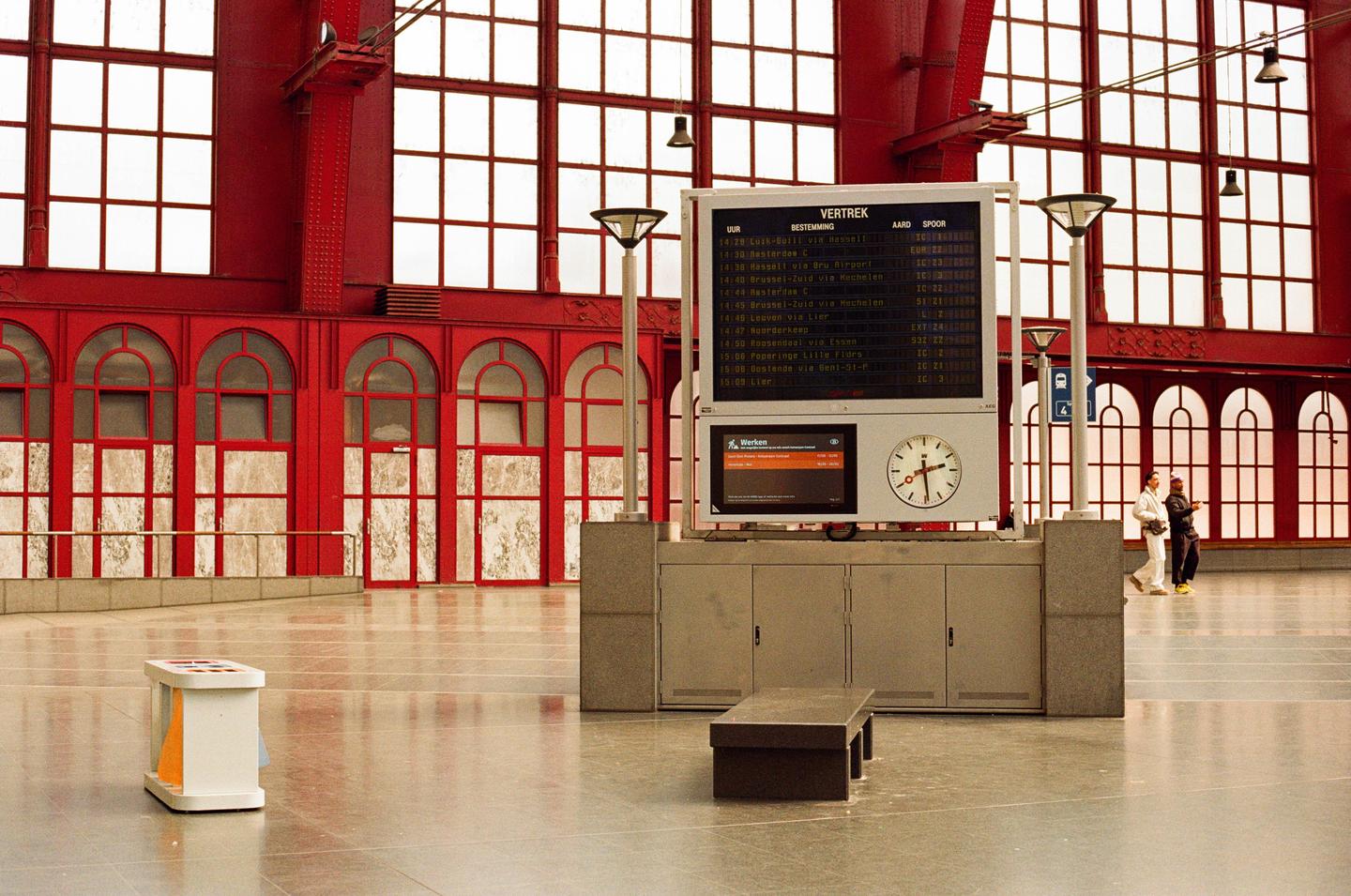 A large red train station interior features a departure board, a clock, and two people walking in the distance.