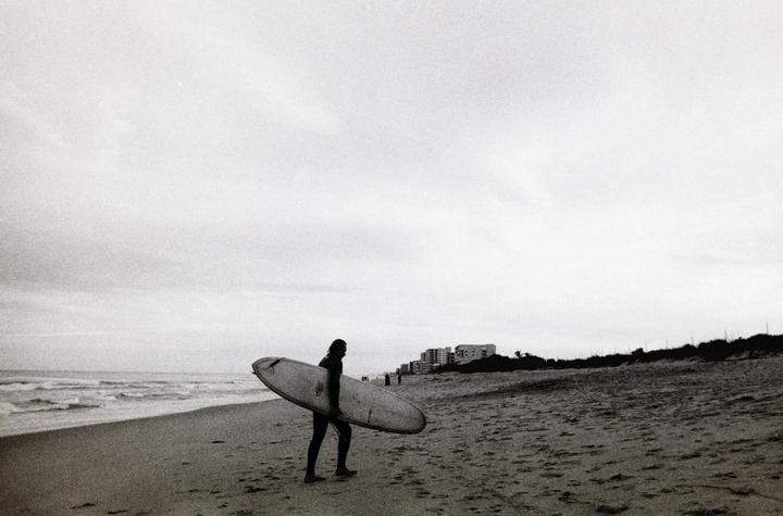 A surfer carrying a surfboard walks along a sandy beach with waves crashing on the left and buildings in the distance on the right.