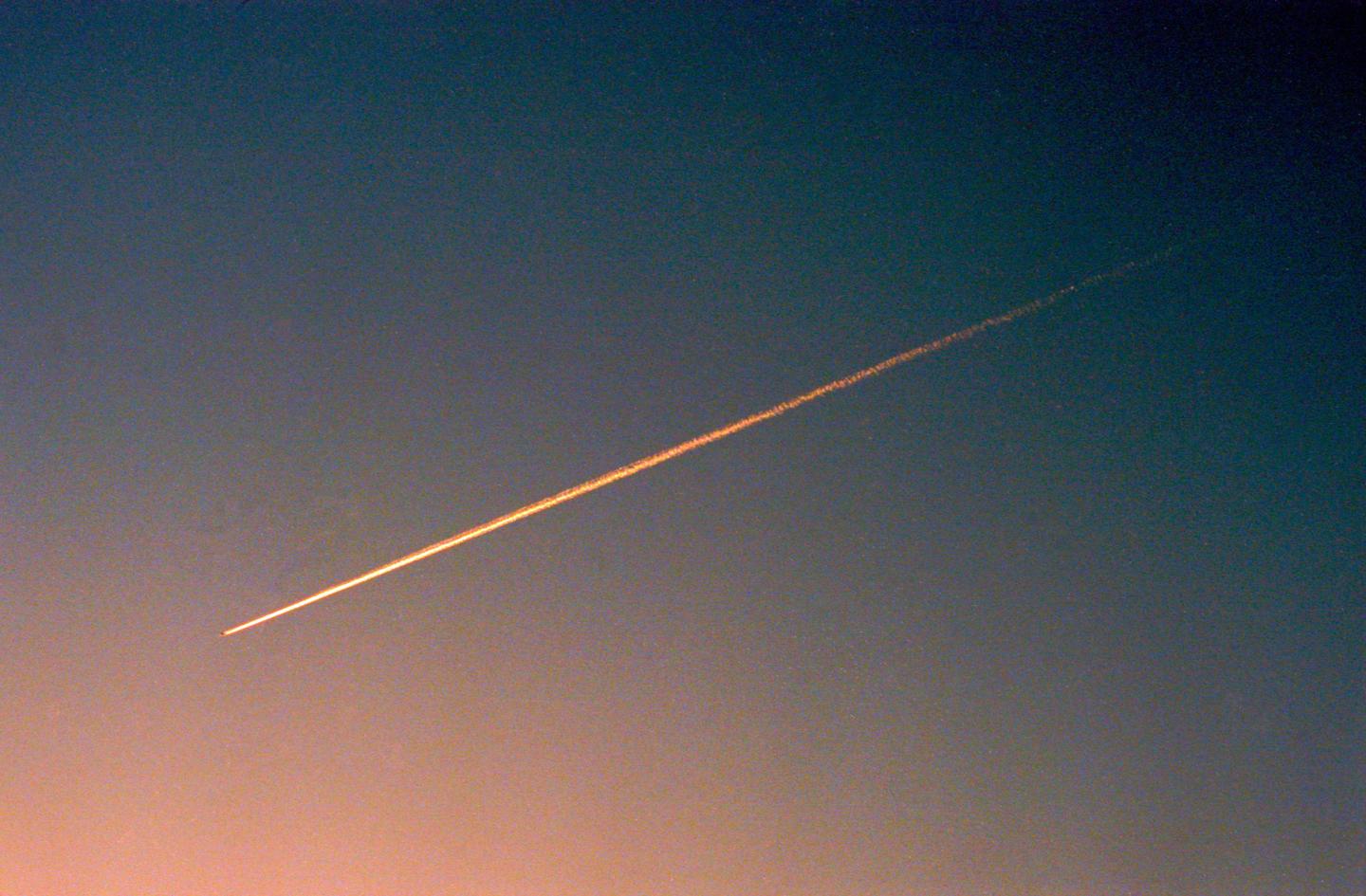 A single contrail streaks across a gradient sky from blue to orange.