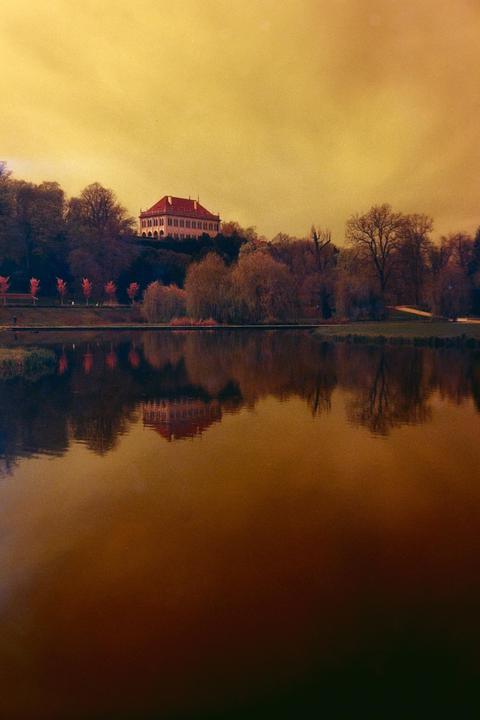 A large building with a red roof sits on a hill overlooking a body of water, reflecting in its surface.