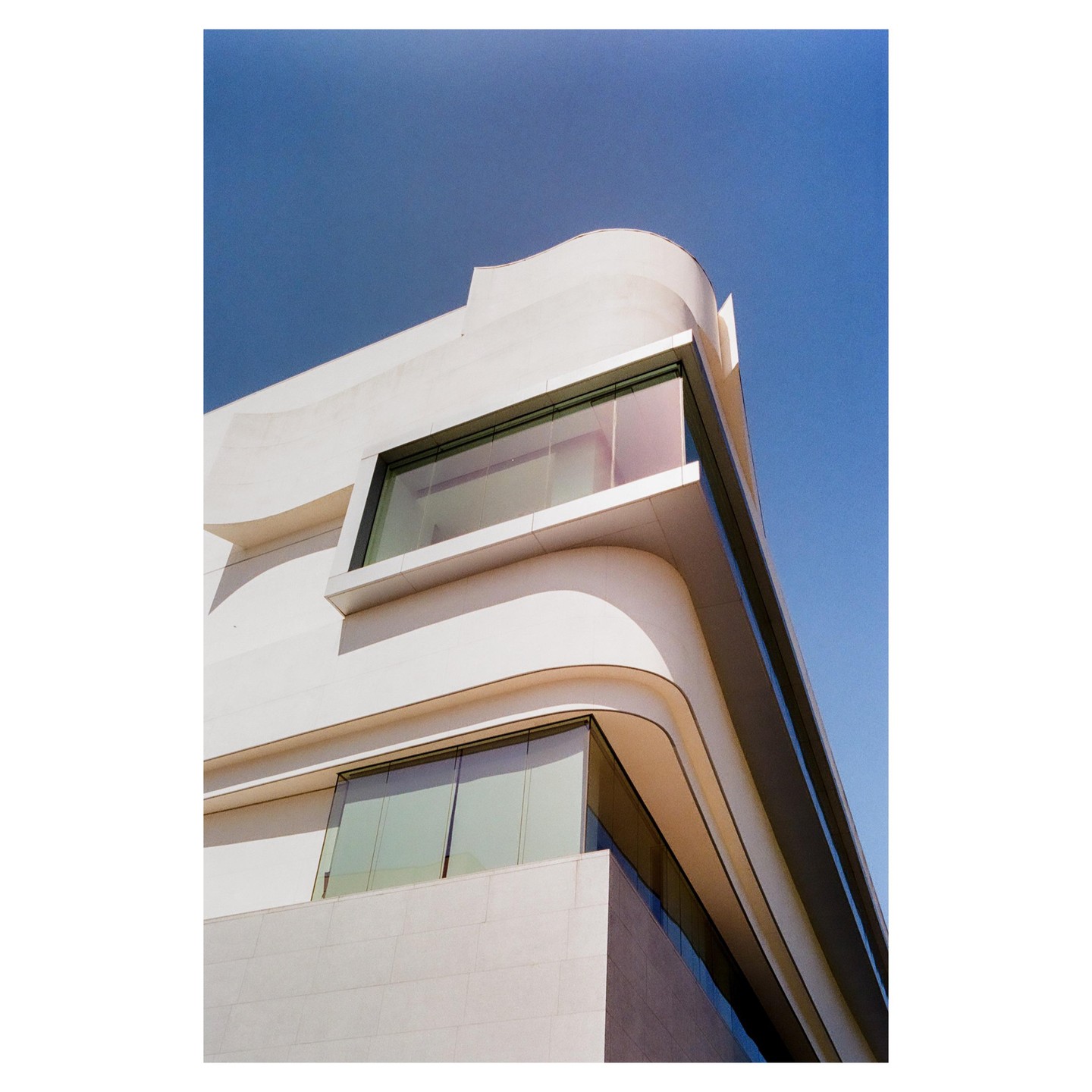 A modern building with curved white walls and large windows is shown against a clear blue sky.