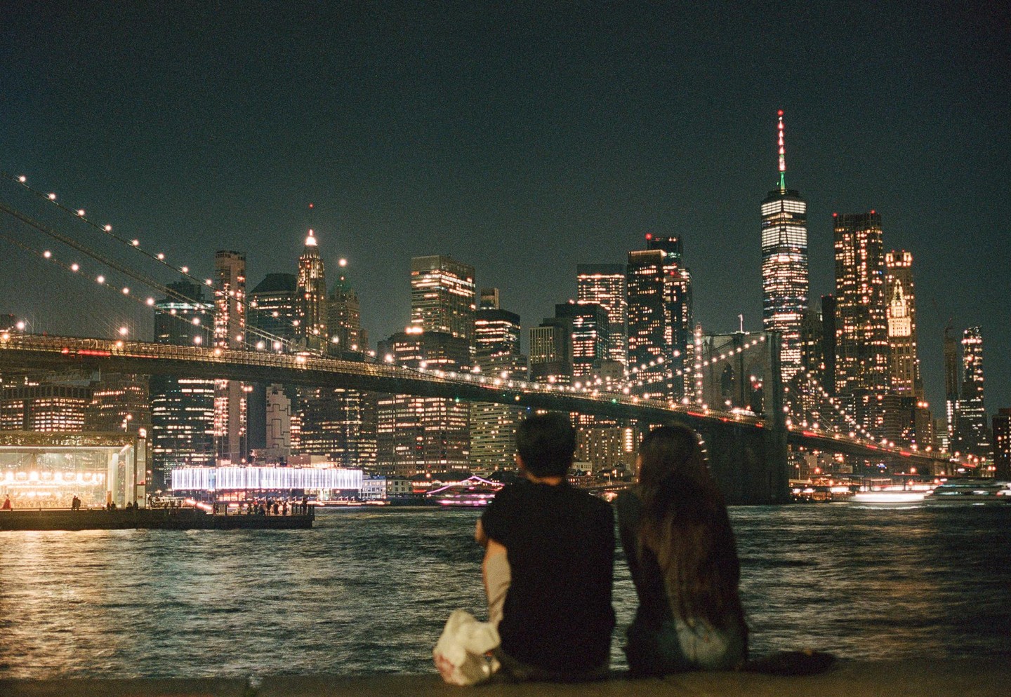 A couple sits by the water at night, looking at the illuminated Brooklyn Bridge and the New York City skyline.