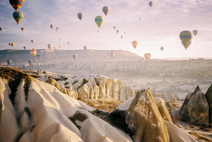 Hot air balloons float above a mountainous landscape with people standing on a ridge.