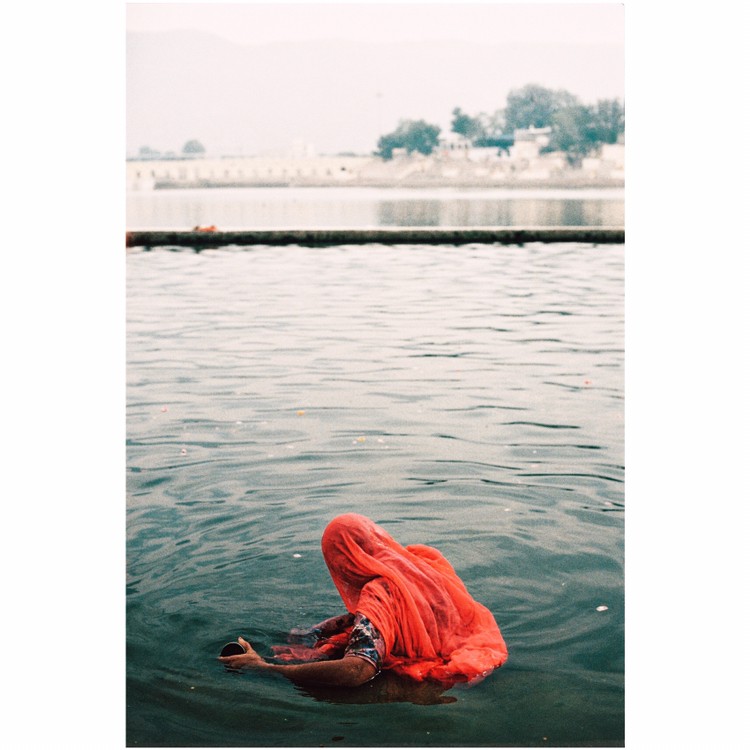 A woman in a red garment is partially submerged in water, holding a small bowl.