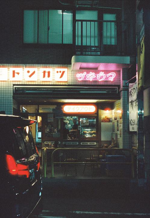 A car is parked in front of a brightly lit storefront with neon signs at night.
