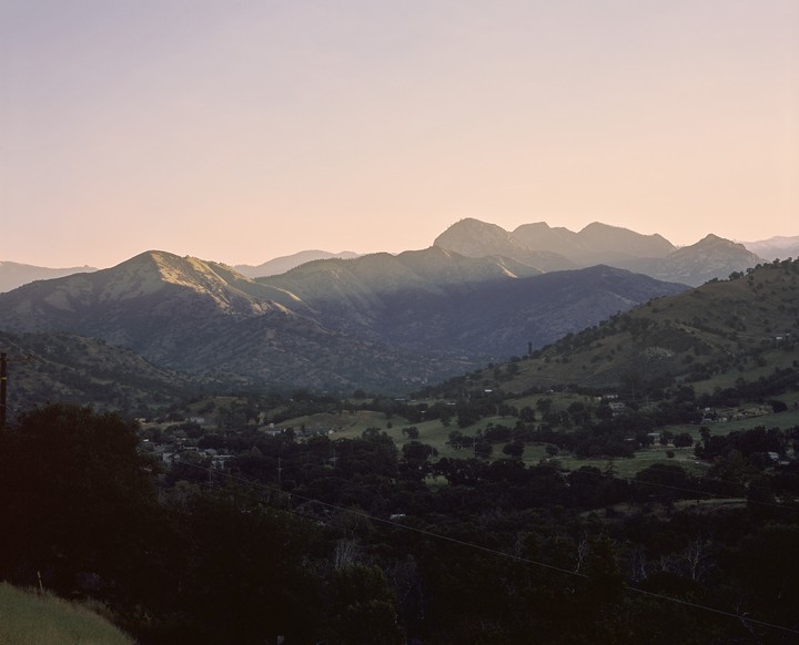 A valley with trees and houses is nestled between mountains under a hazy sky.