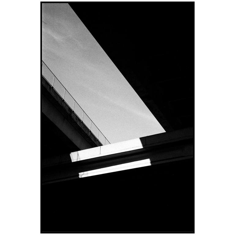 A black and white abstract image shows a diagonal concrete overpass with a railing, and two fluorescent lights below.